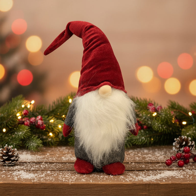 Christmas gnome with red hat and white beard standing on a wooden surface with festive decorations and lights in the background.